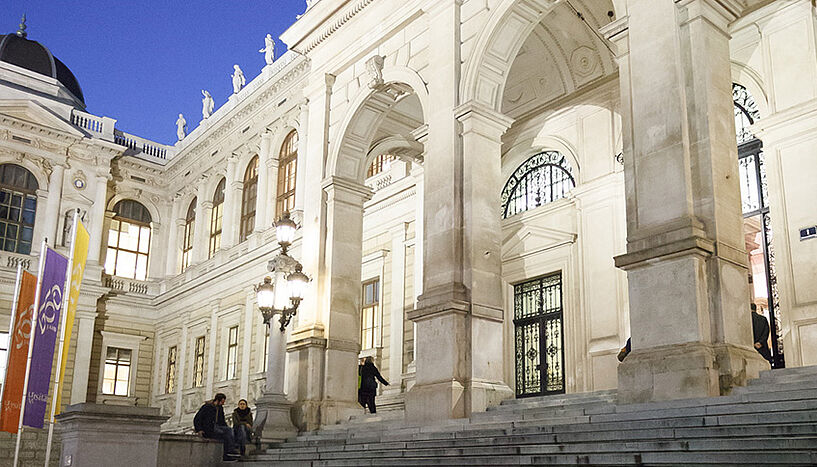 Das Foto zeigt das Hauptgebäude der Universität Wien, festlich beleuchtet am Abend.