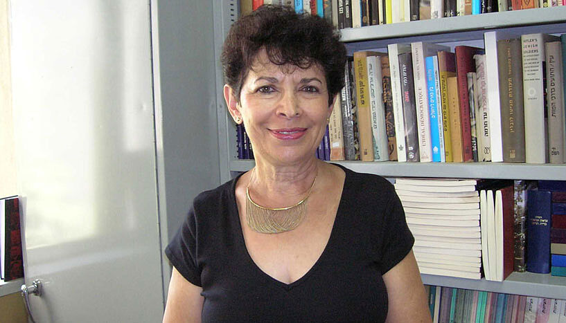 Die israelische Historikerin Dina Porat steht vor ihrem Bücherregal und lächelt in die Kamera.