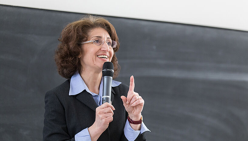 A professor is standing in front of a blackboard, holding a microphone in one hand and explaining something.