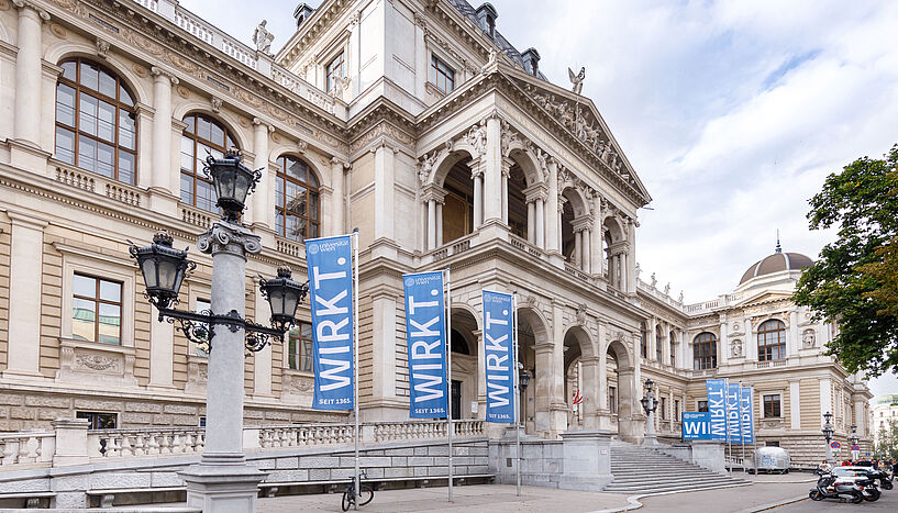Außenansicht des Hauptgebäudes der Universität Wien mit blauen Flaggen, auf denen "Wirkt" steht