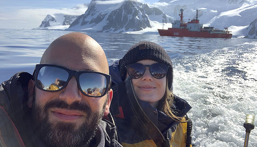 In the foreground two people with sunglasses looking into the camera, surrounded by icy sea. In the background a research vessel and, behind that, snow-covered mountains.