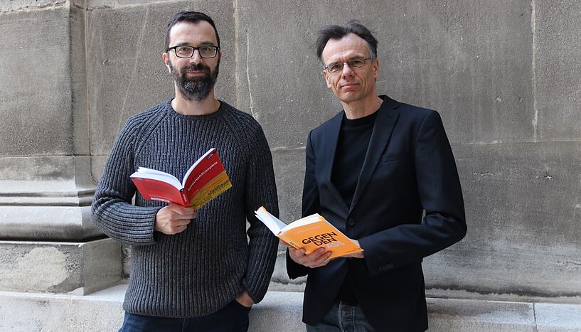 Matthias Flatscher und Gerald Posselt jeweils mit einem Buch in der Hand, vor einer Steinmauer stehend.