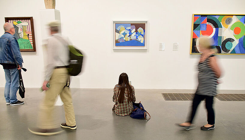 People in a gallery, some standing, some sitting, some walking in front of a wall with abstract paintings.