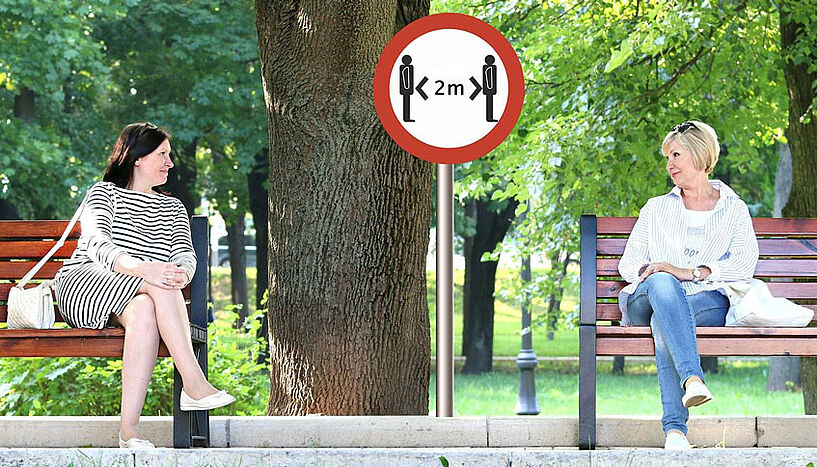 2 Frauen mit zwei Meter Abstand sitzen auf zwei Parkbänken und unterhalten sich