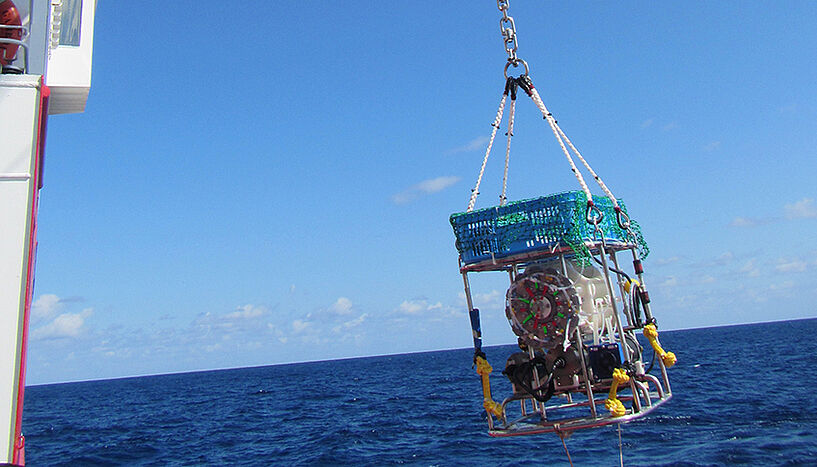 Ein großes Gerät hängt an einem Kran über die Bordwand eines Forschungsschiffes. Im Hintergrund das klare Meer und ein wolkenfreier Himmel.