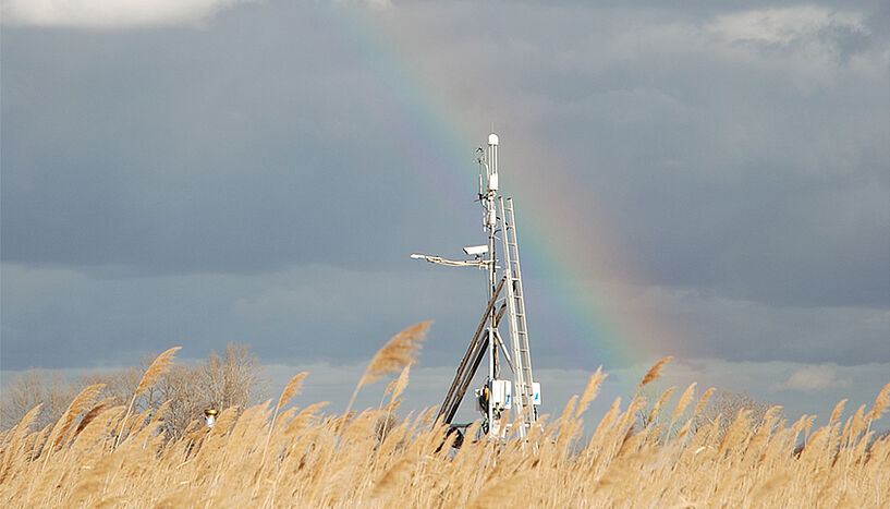 Ein Messturm hinter einem Getreidefelder, in der Ferne ein Regenbogen vor einem Hügel