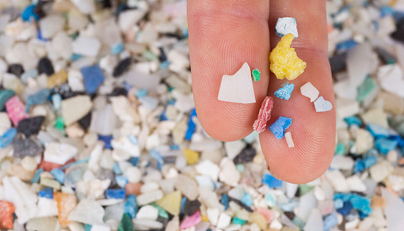 Vor dem Hintergrund eines Plastikberges liegen kleine Plastikteile auf zwei Fingern.
