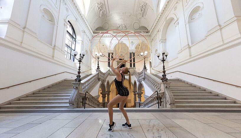 Eine Akrobat*in führt eine Pose mit Reifen im Hauptgebäude der Universität Wien vor. 