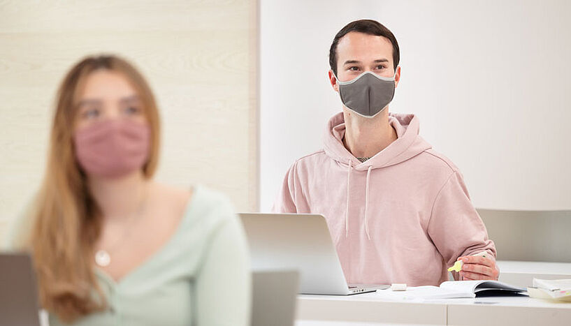 Zwei Studierende nehmen an einer Lehrveranstaltung in einem Seminarraum teil. Sie tragen Masken.