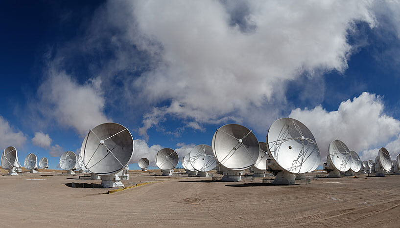 Eine breite Aufnahme der riesigen Radioteleskope bei Tageslicht - die weißen Teleskope stehen auf kargem Untergrund und leuchten vor dem bewölten Himmel hervor.
