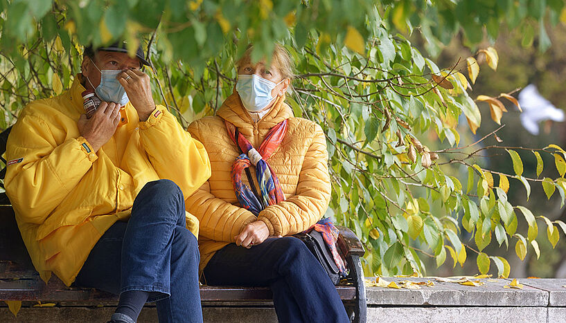 A couple with masks sitting on a park bench underneath a tree.