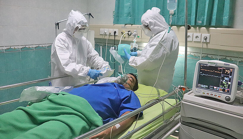 Bild eines Patienten, der in einem Krankenhausbett liegt, und von 2 Ärzten im Schutzanzug beatmet wird 