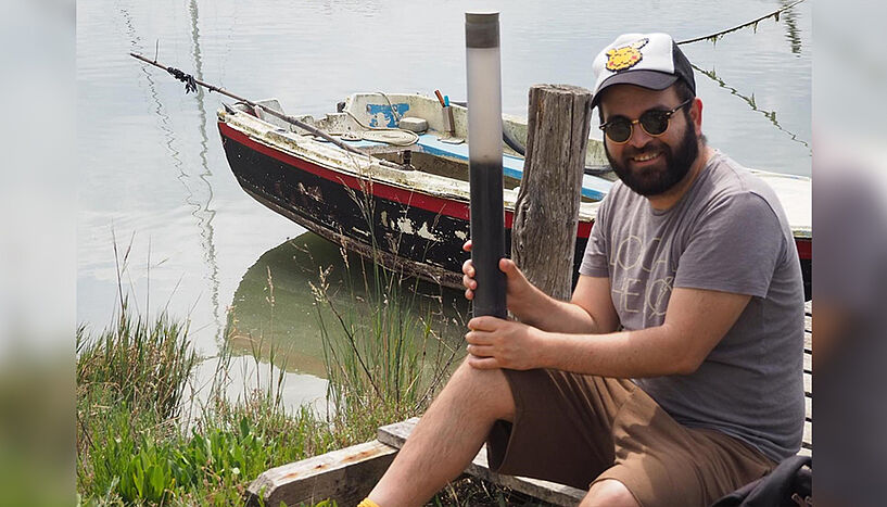 A portrait photograph of Rafael Ponce sitting on a riverbank with a specimen collection tube.