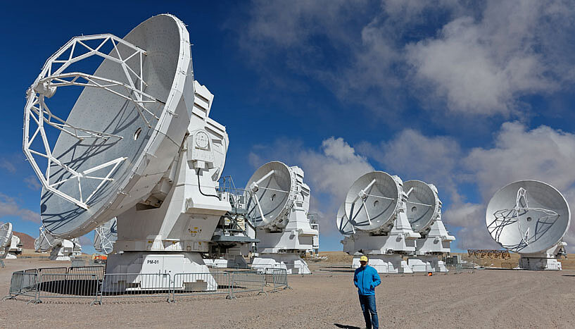 Der Astrophysiker, Franz Kerschbaum, steht mit Schutzhelm neben einem der großen Teleskope in den chilenischen Anden - dadurch wird erkenntlich, wie groß die Teleskope sind. 