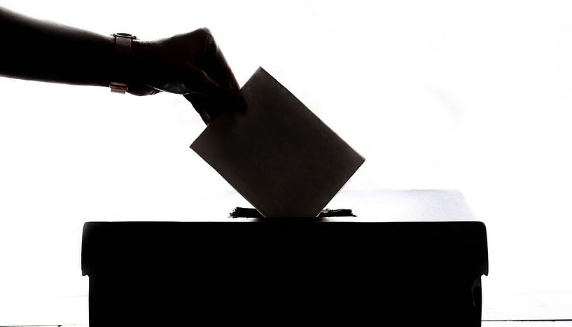 Eine Wahlkarte wird in eine Wahlurne geworfen. 