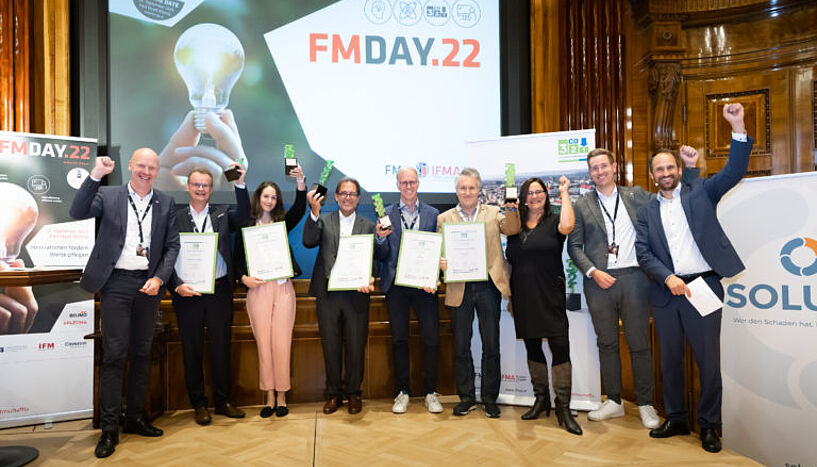 neun Personen stehen in einem Bogen vor einem Transparent des FM Days 22. Die Personen halten ihren Preis, eine grüne Auszeichnung, in der Hand