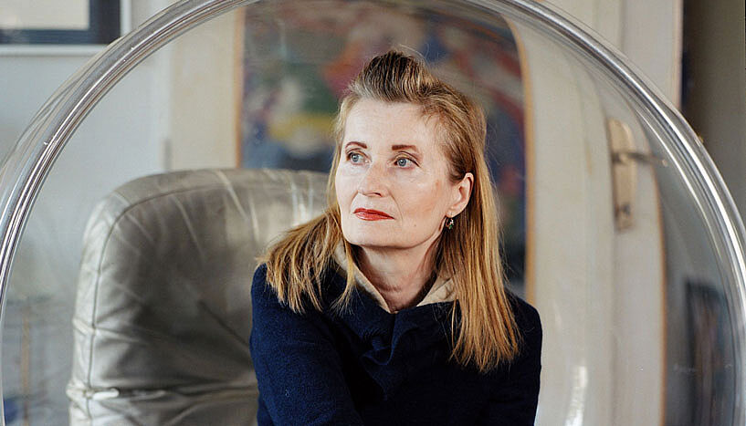 Elfriede Jelinek sitzt mit ernstem, visionären Blick in einem dursichtigen Hängesessel.