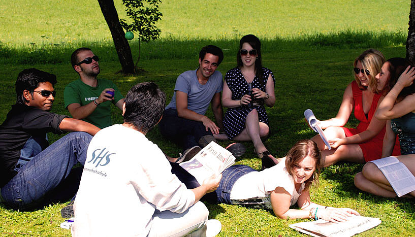 Studierende sitzen im Schatten eines Baumes auf einer Wiese und tauschen sich zu Studieninhalten aus.