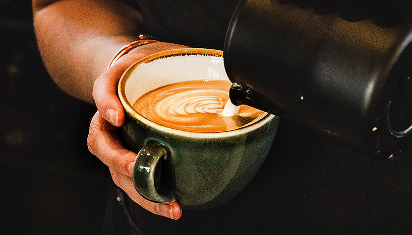 A hand is holding a coffee cup into which milk foam is being poured.